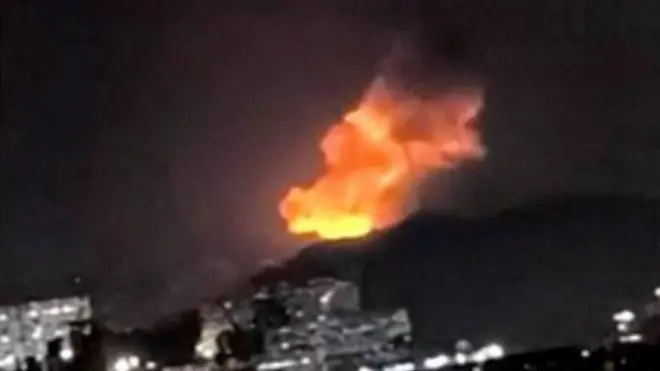 粗い画像。夜空を背に黒いシルエットになっている山の稜線の向こうで、オレンジ色の炎と煙が上っている。手前に市街地が広がる
