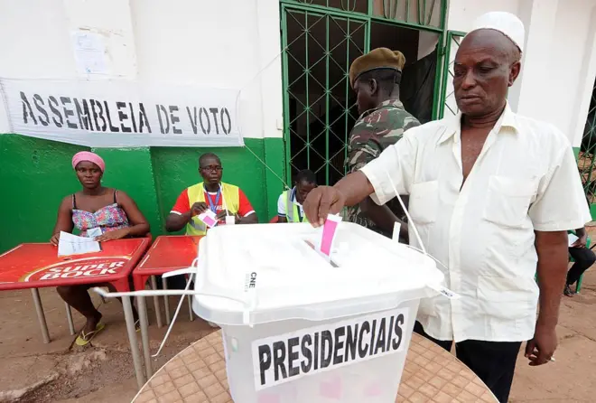 Un homme vêtu d'une chemise blanche avec un chapeau blanc met un bulletin dans une urne transparente sur laquelle est écrit en portugais "Presidenciais", un homme en treillis debout et trois personnes assis autour d'une table sont visible en arrière-plan.