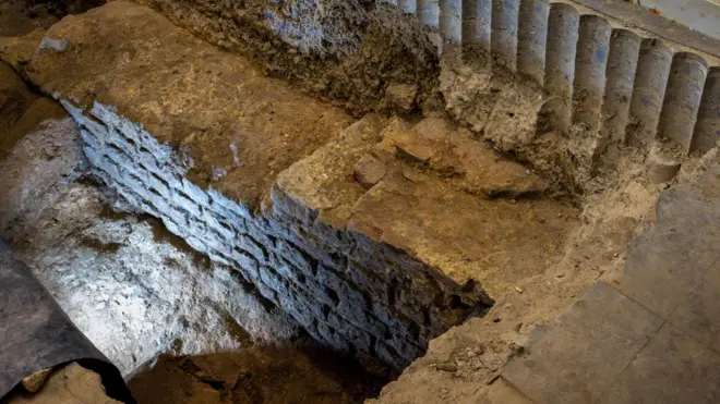 Uma parte do muro de pedra com cerca de três metros de comprimento, ao fundo de uma vala cavada no porão de um prédio, junto a estantes e prateleiras