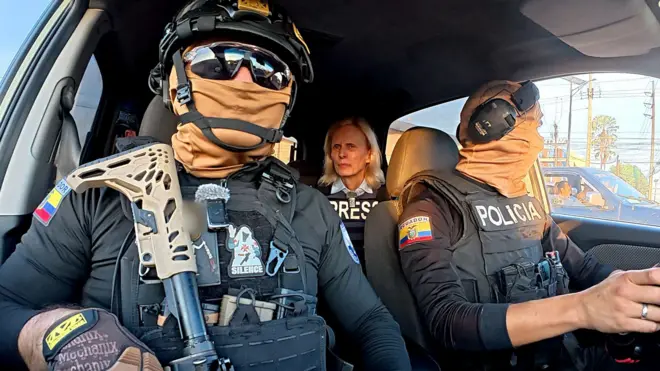 Two Ecuadoran police officers in dark sunglasses, helmets and balaclavas - which entirely obscure their faces - sit in a car in front of the BBC's Orla Guerin, wearing a flak jacket with the word "PRESS" on it.