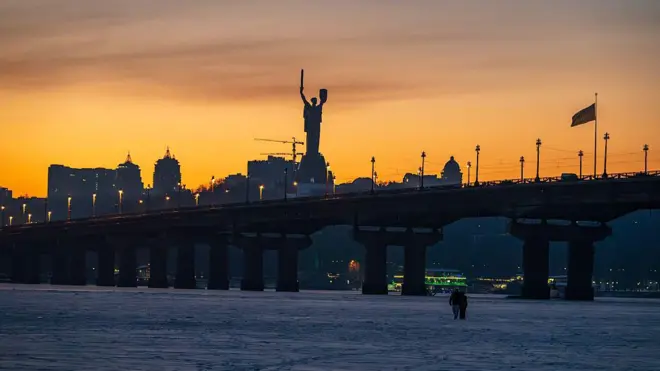 Зимовий краєвид Києва під час заходу сонця. На передньому плані – вкрита льодом поверхня Дніпра, по якій ідуть двоє людей. Над ними проходить міст, уздовж якого тягнеться лінія ліхтарів. На горизонті видно темний силует міських будівель та велику фігуру Батьківщини-матері.