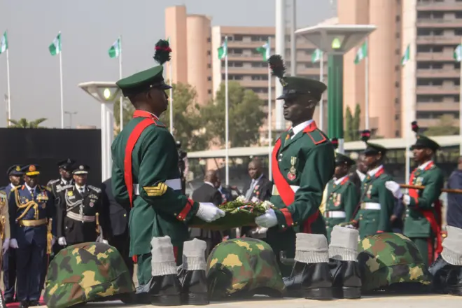 Àwọn ológun méjì tó dúró wámú, tí fìlà àti bàtà àwọn ológun wà ní ẹ̀gbẹ́ wọn. Àwọn ológun púpọ̀ wà lẹ́yìn wọn