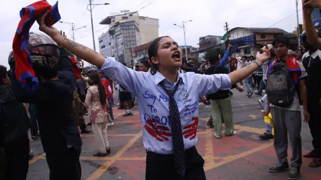 A young woman, wearing what resembles a school uniform which has been emblazoned with protest slogans, shouts and holds her arms and the Nepali flag in the air