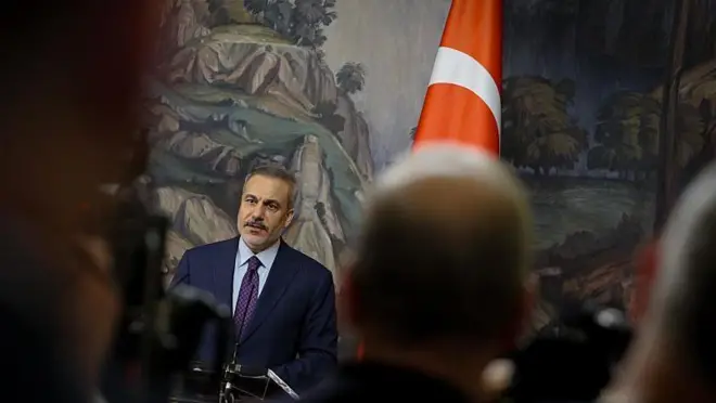 Hakan Fidan, gazetecilerin kafalarının arasından çekilen fotoğrafta yalnız başına konuşurken görülüyor. Yanında Türk bayrağı var.