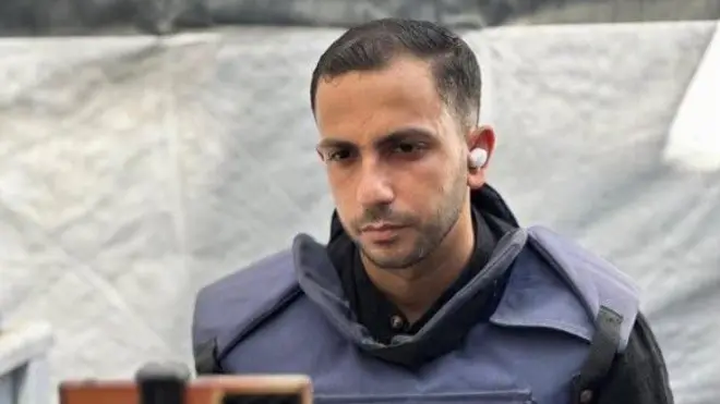 أنس الشريف يرتدي سترة الصحفيين وينظر إلى هاتفه الخلوي المثبت أمامه، ويبدو أنه يجهز لتغطية إخبارية.