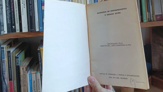Imagem de uma mão segurando cópia do Glossário de Entorpecentes e Drogas Afins, da Polícia Federal, com uma prateleira de livros atrás