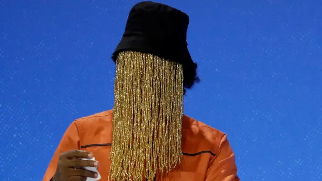 Anas Aremeyaw Anas dey speak on stage for press freedom event in London. E wear black hat and im face dey covered by long columns of yellow beads.