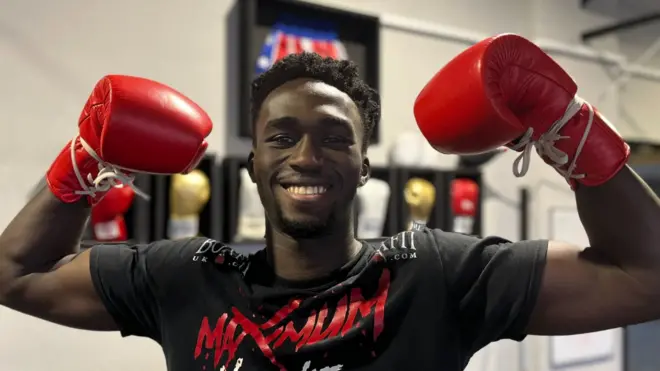 Adam Olaore smile as e hold e arms up for victory pose while e wear red boxing gloves