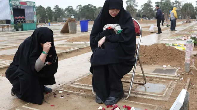 墓の前で黒い布で全身を覆った1人がしゃがみ、もう1人がパイプ椅子に腰かけて下を見ている。少し離れたところに他の人たちもいる