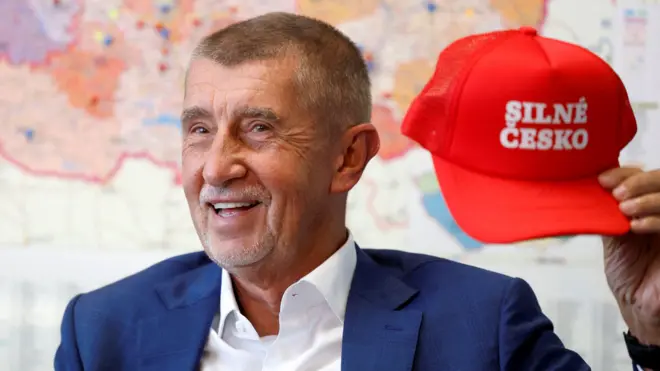 Andrej Babisz w białej koszuli i niebieskiej marynarce trzyma czerwoną czapkę baseballową z hasłem „Silne Czechy”. W tle na ścianie wisi mapa.