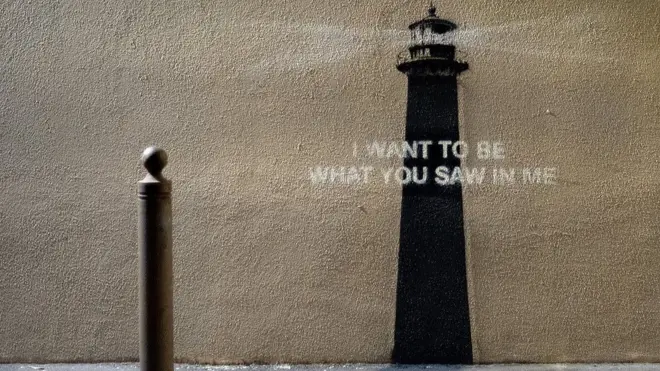 A photo of the Banksy piece of art. A metal bollard is in the foreground on the pavement, while a painting of a lighthouse is on the wall superimposed with a quote "I want to be what you saw in me". 