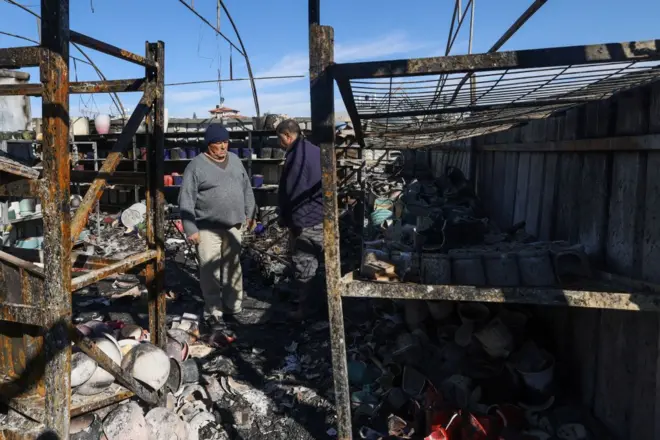 رجلان يرتديان ملابس شتوية يقفون في مكان متضرر من الهجمات. الصورة التقطت نهاراً.