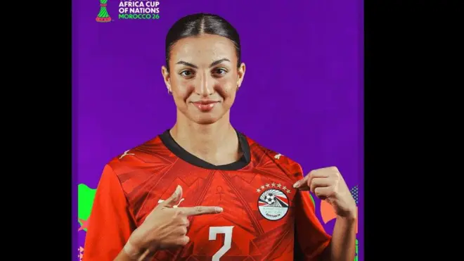 لاعبة لون بشرتها حنطي وترتدي قميص المنتخب المصري للرجال باللون الأحمر، وعليه شعار الاتحاد المصري وفوقه سبع نجوم.