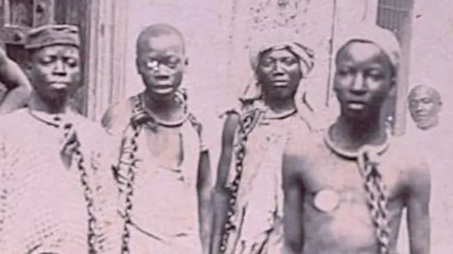 A 19th Century photograph shows black enslaved people in chains at a market on the island of Zanzibar. They can be seen looking at the camera.