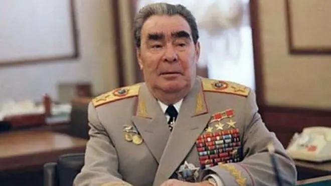Похилого віку Брежнєв сидить за столом у кабінеті, одягнений у світлий військовий мундир із темною краваткою та численними медалями, орденами та кольоровими планками; на плечах видно оздоблені погони.
На тлі — розмитий інтер’єр із шторами, меблями та телефоном праворуч.