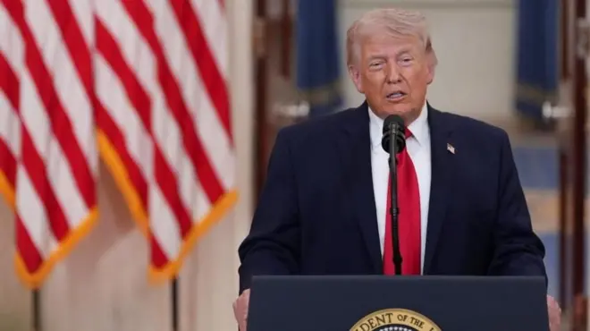 Trump koyu renk ceketi ve kırmızı kravatı ile başkanlık kürsüsünde mikrofon önünde konuşuyor, arkasında Amerikan bayrağı var