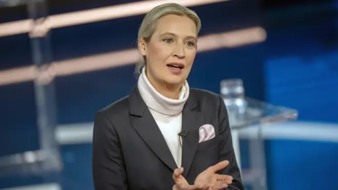 أليس فايدل، مرشحة حزب البديل من أجل ألمانيا، بيضاء البشرة شعرها أشقر، ترتدي قميصاً شتوياً أبيض ياقته عالية وعليه جاكيت رسمي لونه رمادي