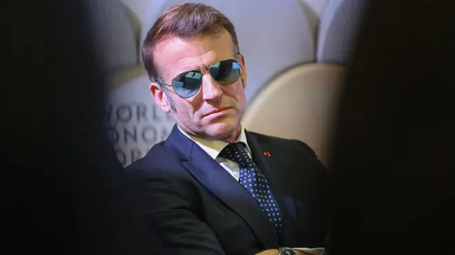 Fransa Cumhurbaşkanı Macron zirveye güneş gözlüğü takıyor. Fotoğrafta ellerini kavuşturmuş ve somurtuyor.