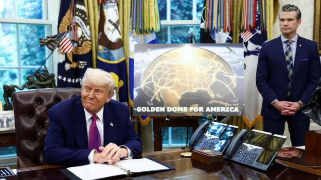 US President Donald Trump is seated at his desk in the White House Oval Office. Stood beside a poster of the Golden Dome is US Defence Secretary Pete Hegseth.