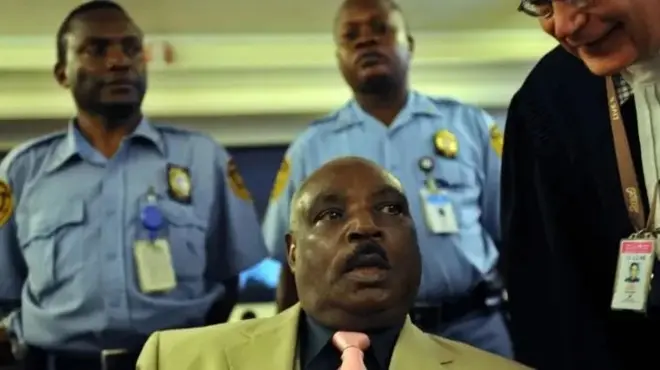 Protais Zigiranyirazo (imbere hagati), yambaye ikaruvari, ishati yirabura n'ikoti ishaka gusa n'umuhongo. Inyuma yiwe hahagaze abapolisi ba Onu babiri bambaye ishati zisa n'ubururu, bambaye n'ibibaranga ku mifuko y'amashati. Ku ruhande rw'i buryo, hahagaze uwumuburanira yambaye ikanzu yirabura