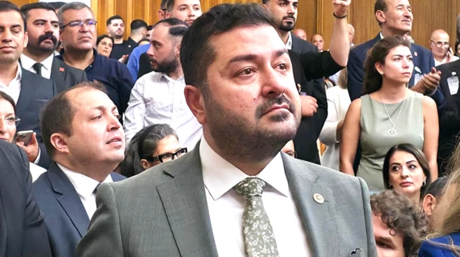 Sakallı bir erkek kalabalık bir ortamda bir konuşmayı dinliyor. Arkada kadınlar ve erkeklerden oluşan bir kalabalık var. En önde o görünüyor.