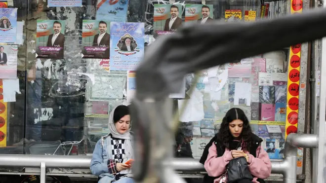 زنی بدون حجاب در کنار زنی با روسری عقب رفته در ایستگاه اتوبوسی نشسته‌اند و مردی در کنار آنها ایستاده است و تبلیغات انتخاباتی پشت آنها دیده می‌شود