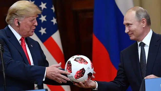 Vladimir Putin smiles as he hands a white and red football to Donald Trump during a meeting in Helsinki, Finland, in July 2018. Behind the pair are a US and a Russian flag. 
