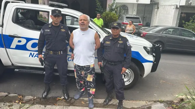 El abogado constitucionalista Enrique Anaya con los brazos hacia atrás y esposado, escoltado por dos policías delante de una camioneta policial.