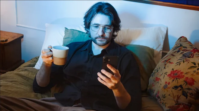 ہاتھ میں کپ پکڑے ایک شخص موبائل فون دیکھ رہا ہے۔ اس کے چہرے پر نیلی روشنی پڑ رہی ہے