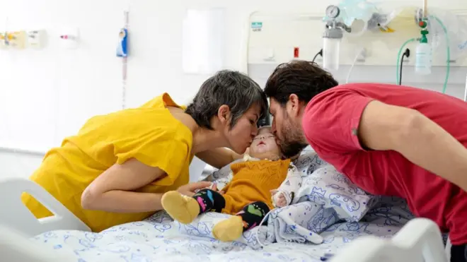 Foto de um bebê em uma cama de hospital com um homem beijando sua bochecha de um lado, e uma mulher, do outro. 