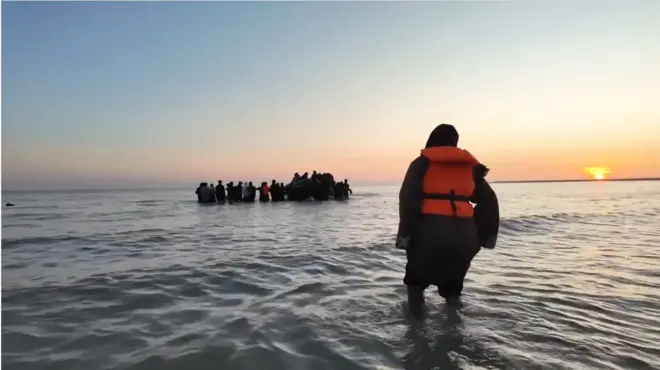 フランスの海岸の浅瀬から、イギリスに向かうゴムボートに乗ろうとする人々。オレンジ色の救命胴衣を着けた1人が、ボートに向かって歩いている。水平線には太陽がかかっている