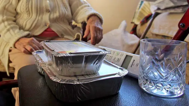 Two foil food containers stacked, and empty glass and a landline wireless phone sit on a black leather-like surface. Behind it is an elderly person wearing a beige cable knit cardigan with buttons. Face is not visible and clothing does not make it possible to distinguish if they are male or female.