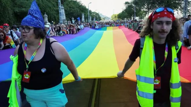 Uczestnicy parady niosą tęczową flagę o długości kilkudziesięciu metrów. Na pierwszym planie widoczne dwie osoby trzymające przód flagi. Jedna z nich ma na sobie czarny T-shirt i żółtą odblaskową kamizelkę. Druga - ubrana w czarną koszulkę na ramiączkach - trzyma odblaskową kamizelkę pod pachą.