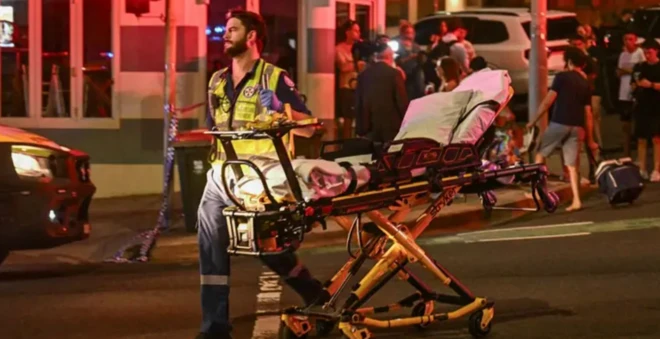 Socorrista empurra uma maca vazia em uma rua à noite, em praia no sul da Austrália, com luzes de emergência ao fundo e pessoas reunidas perto de viaturas policiais.