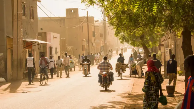Une route sur laquelle roulent des motocyclistes, avec des arbres sur le côté droit et des bâtiment à gauche.