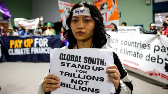 ناشط يحمل لافتة أثناء احتجاج للمطالبة بتمويل مناخي طموح والتخلص التدريجي من الوقود الأحفوري في المنطقة الزرقاء خلال مؤتمر الأمم المتحدة لتغير المناخ COP29 - مكتوب على اللافتة: "الجنوب العالمي: قفوا من أجل التريليونات لا المليارات".