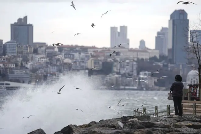 İstanbul Üsküdar'da lodos etkisiyle deniz dalgası kıyıya vurmuş ve martılar havada uçuşuyor, bir kadın kıyıda duruyor, arka planda apartmanlar görünüyor
