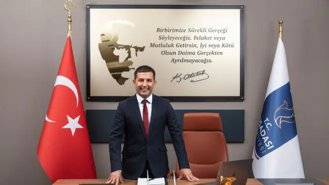 Ömer Günel makamında gülümseyerek poz veriyor. İki yanında Türk bayrağı ve Kuşadası Belediyesi bayrağı var. Arkasında ise Atatürk'ten çerçeveli bir alıntı görünüyor.