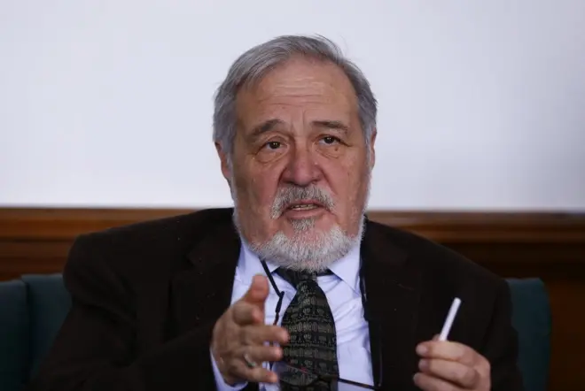 İlber Ortaylı, bir elini ileriye doğru kaldırmış, diğer elinde kalem var. Konuşuyor