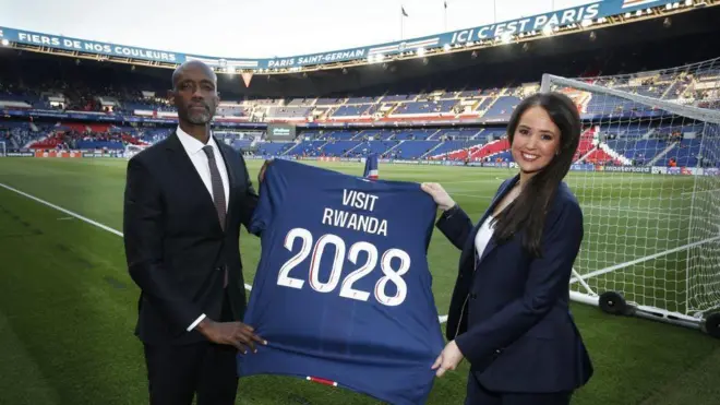Umuyobozi w'ikigo cy'u Rwanda cy'iterambere Jean-Guy Afrika (ibumoso) hamwe n'umugore wo mu buyobozi bwa PSG, bafashe umupira w'amaboko magufi wa PSG wanditseho 'VISIT RWANDA 2028'