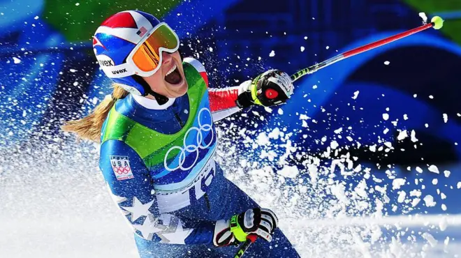 Лыжница радуется успеху, подняв в небо сотни снежных "брызг". На ней форма с олимпийскими кольцами