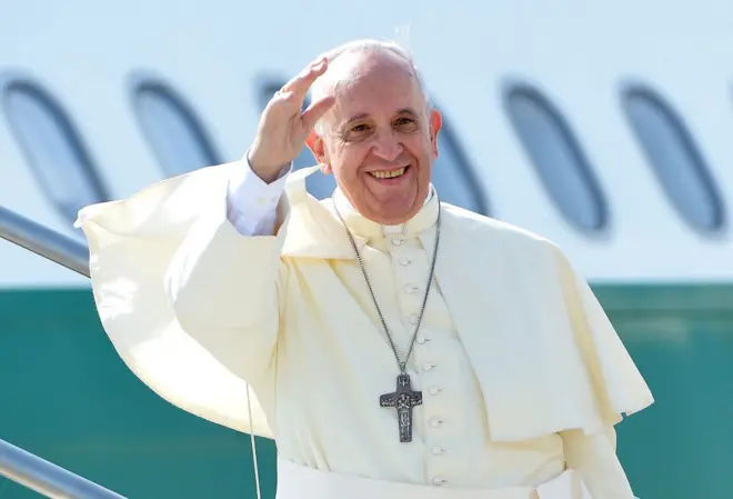 Pape François dans une robe blanche avec une croix au cou, lève la main droite en signe de salutation et sourit