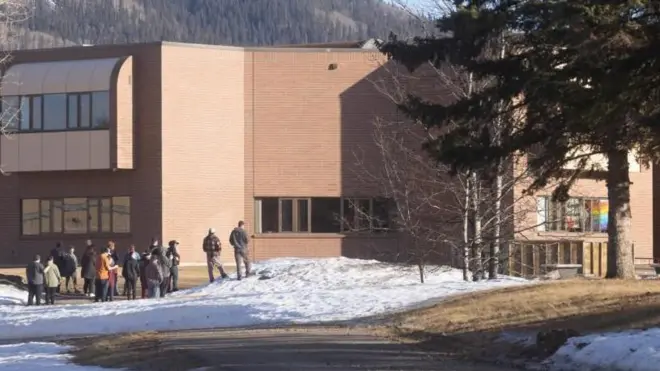 晴れた日に、山を背に明るい茶色の建物がたち、人が何人かその前に集まっている。地面にはとけかかった雪が見える