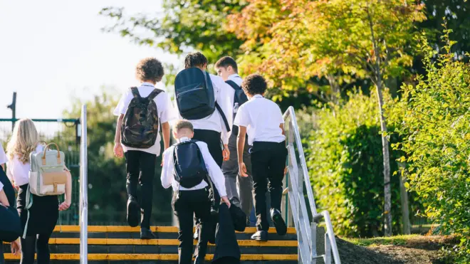 Beyaz gömlekli ve koyu renk pantolonlu öğrenciler sırt çantaları ile arkaları dönük halde merdivenden çıkıyor