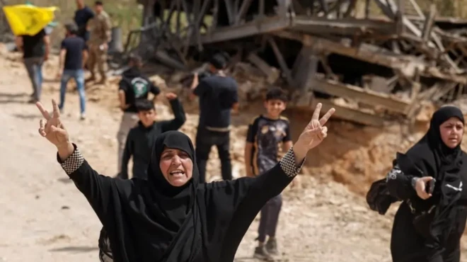 دو زن محجبه با لباس سیاه رنگ، یکی از آنها دستانش را بالا برده وعلامت پیروزی نشان می‌دهد، در پسزمینه ساختمانی ویران و چند کودک دیده می‌شوند