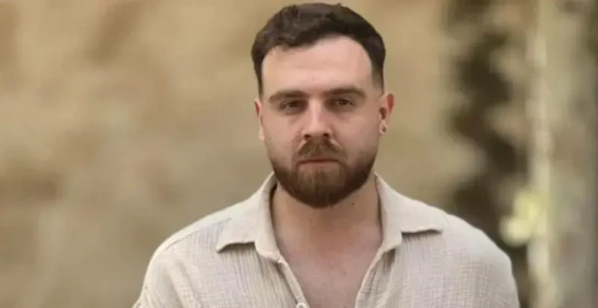 Gömlekli kumral bir erkek kameraya bakıyor. Gömleğinin düğmeleri açık ve genç bir görüntüsü var. Kulağında da küpesi dikkat çekiyor.