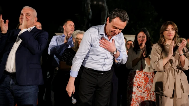Muškarac u košulji se poklanja publici, iza njega ljudi, u ruci drži mikrofon