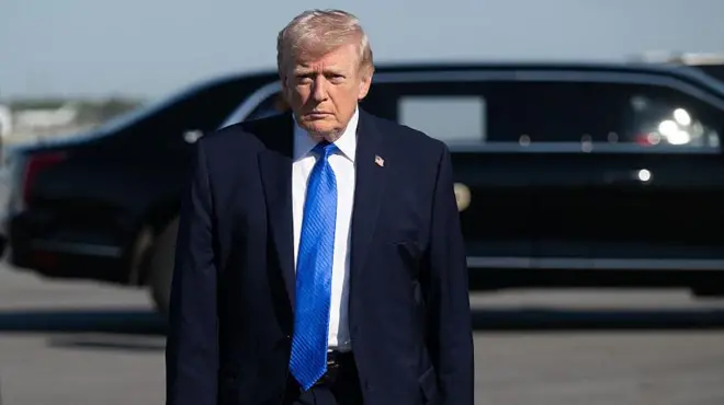 دونالد ترامپ مقابل دوربین عکاسی ایستاده و مستقیم به آن نگاه می‌کند یک خودروی مخصوص او در پشت سر او دیده می‌شود