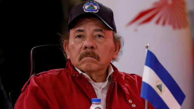 Daniel Ortega, de boné e jaqueta vermelha, ao lado de uma pequena bandeira da Nicarágua