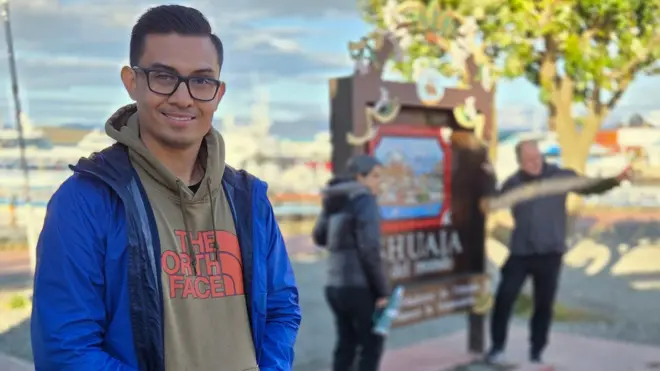Hombre sonríe a la cámara mientras dos personas detrás de él se toman una foto